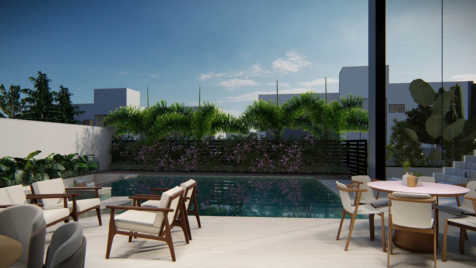 Wide paved terrace with wooden armchairs and a round dining table beside the pool, backed by a green wall of palms and flowering groundcover.