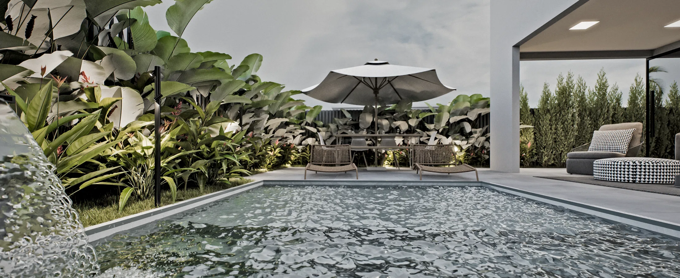Rectangular pool with a wall water blade, a border of tropical plants, an umbrella with a table and a covered lounge with a sofa on the right.