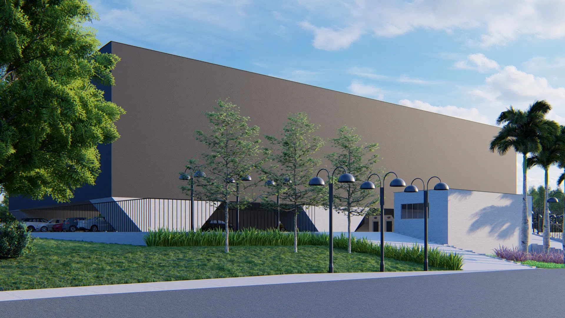 Front façade of the beige warehouse volume, with a dark base, a row of trees, street lamps, lawn and palm trees under a blue sky.