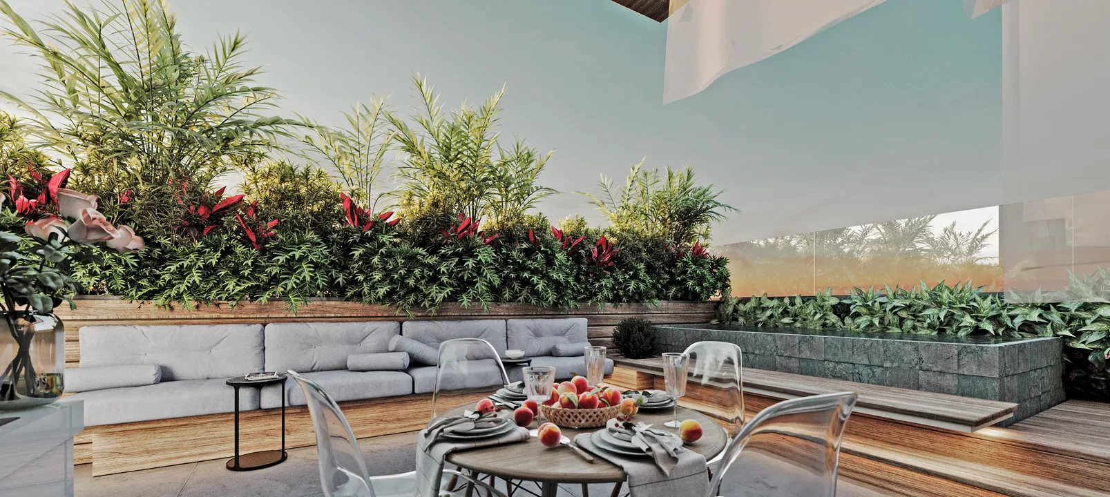 Terrace at dusk with a grey padded sofa beside a wooden planter of foliage and red cordyline, and a round table set with a fruit bowl.